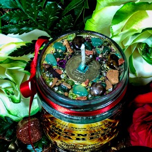 Puede incluir: Una vela de vidrio en un frasco con una cinta roja y una etiqueta dorada que dice "Money Maker" con el texto "Ritually Blessed, Fixed & Loaded Prosperity Candle". La vela est&aacute; rodeada de hojas verdes, rosas blancas y rojas, y monedas de oro.
