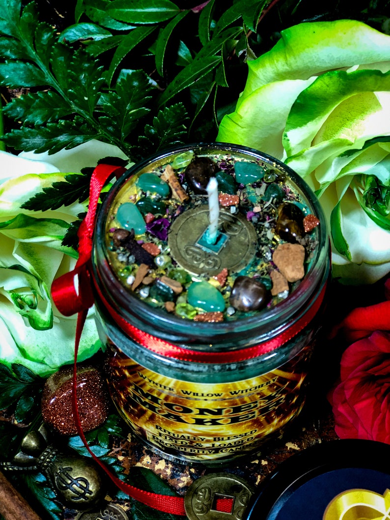 Puede incluir: Una vela de vidrio en un frasco con una cinta roja y una etiqueta dorada que dice "Money Maker" con el texto "Ritually Blessed, Fixed & Loaded Prosperity Candle". La vela est&aacute; rodeada de hojas verdes, rosas blancas y rojas, y monedas de oro.