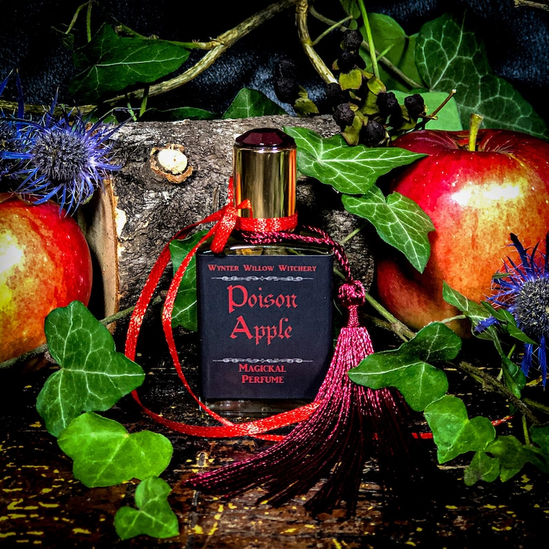 May include: A small bottle of perfume with a black label that reads "Poison Apple" and "Magickal Perfume". The bottle is surrounded by red ribbon, a red tassel, and green leaves.