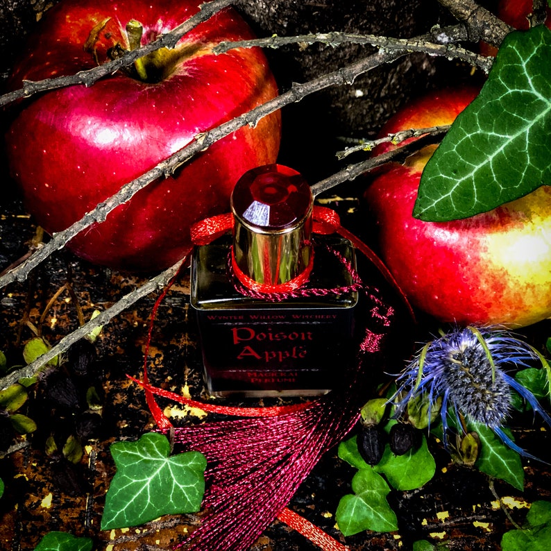 May include: A dark red glass bottle of Poison Apple perfume with a gold cap and a red tassel. The bottle is sitting on a wooden surface with red apples and green leaves.