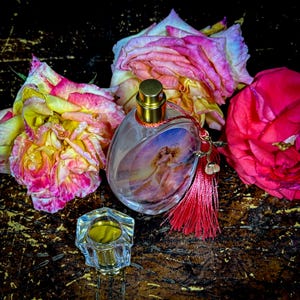Puede incluir: Un frasco de perfume ovalado con tapa dorada y borla roja, con una imagen pintada de una mujer. El frasco est&aacute; rodeado de rosas rosas y amarillas, con un peque&ntilde;o recipiente hexagonal transparente en primer plano.