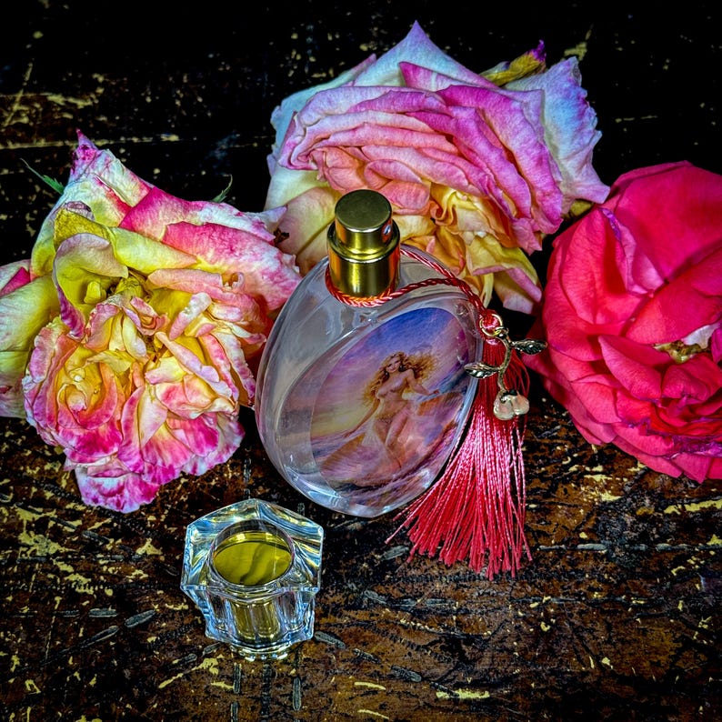 Puede incluir: Un frasco de perfume ovalado con tapa dorada y borla roja, con una imagen pintada de una mujer. El frasco est&aacute; rodeado de rosas rosas y amarillas, con un peque&ntilde;o recipiente hexagonal transparente en primer plano.