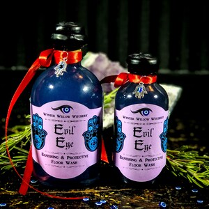 May include: Two bottles of purple floor wash with a label that reads "Evil Eye Banishing & Protective Floor Wash". The bottles are decorated with a red ribbon and a silver charm with a blue evil eye.