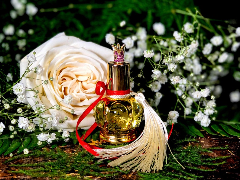 May include: A gold perfume bottle with a crown-shaped stopper and a white tassel. The bottle is surrounded by white flowers and a white rose.