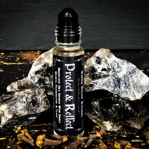 May include: A clear glass roller bottle with a black cap and label that reads "Protect & Reflect" with the text "WYNTER WILLOW WITCHERY" above it. The label also includes the text "PROTECTIVE OILS INFUSED WITH SMOKY QUARTZ, FRANKINCENSE & CLOVE". The bottle is sitting on a surface with several gray stones and brown cloves.