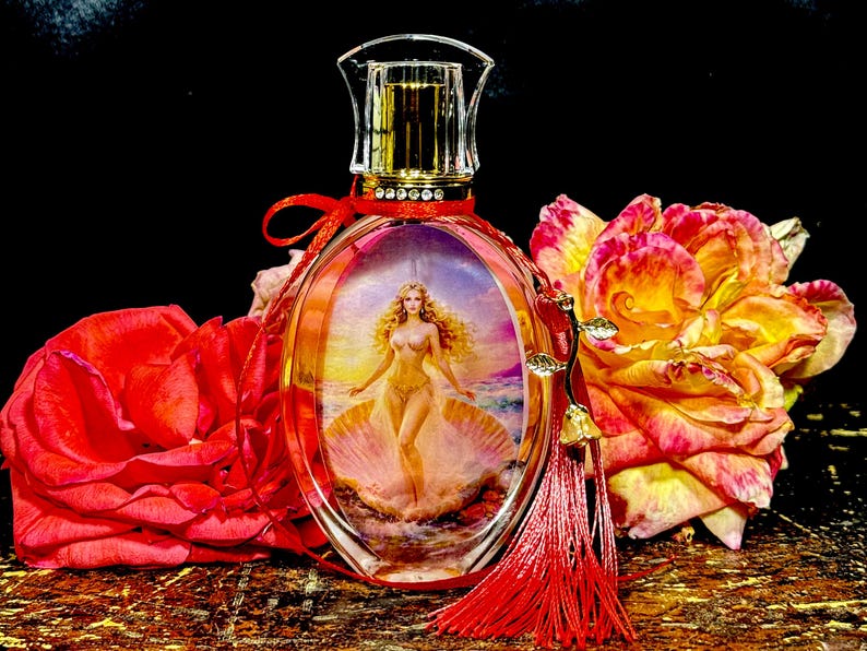 Puede incluir: Frasco de perfume ovalado con tapa de cristal transparente y borde dorado. La botella presenta una imagen de una mujer en una concha. Una cinta roja y una borla adornan la botella. Rosas rojas y amarillas en el fondo.