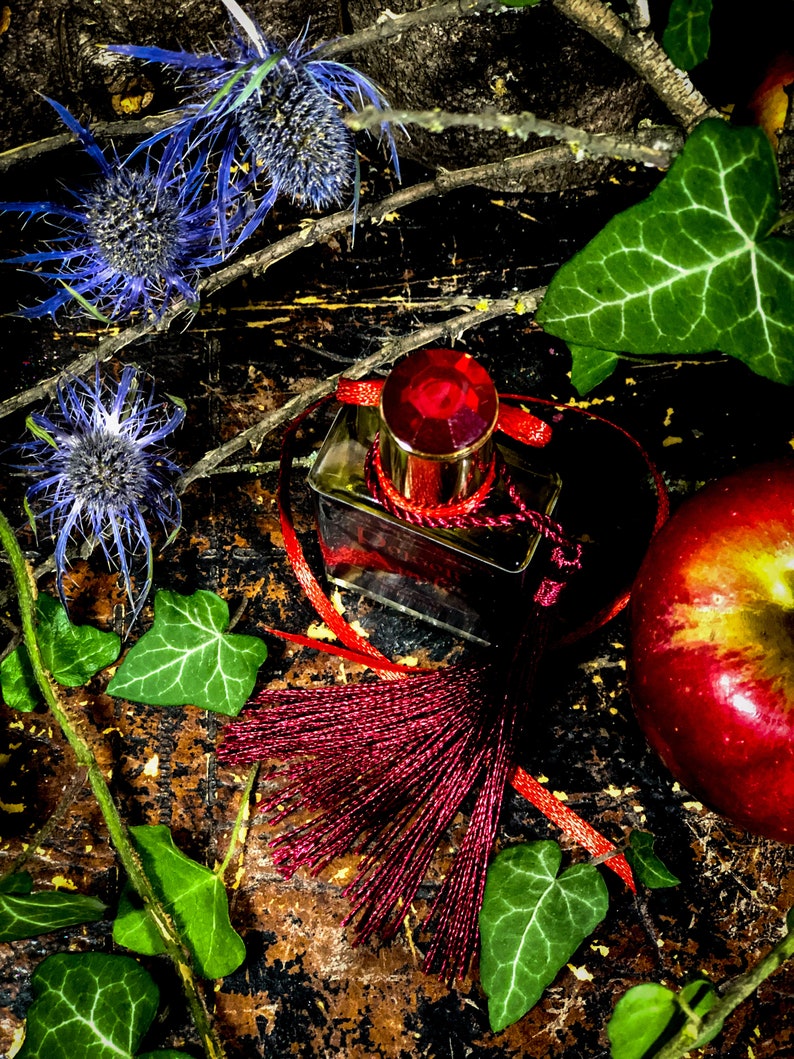 May include: A dark red glass bottle with a red tassel and a red jewel-toned stopper. The bottle is sitting on a wooden surface with green leaves and a red apple.