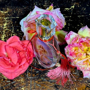 Puede incluir: Un frasco de perfume transparente con un tap&oacute;n decorativo, con una imagen pintada de una mujer. La botella est&aacute; adornada con una cinta roja y una borla, y rodeada de rosas rosas y amarillas. La botella tiene un cuello dorado.