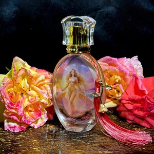 Puede incluir: Un frasco de perfume ovalado con un cuerpo de vidrio transparente, que presenta una pintura de una mujer en una concha. El frasco tiene un cuello dorado y un tap&oacute;n transparente facetado. Una borla roja con detalles dorados cuelga del frasco. Rosas en tonos rosa y amarillo rodean el frasco.