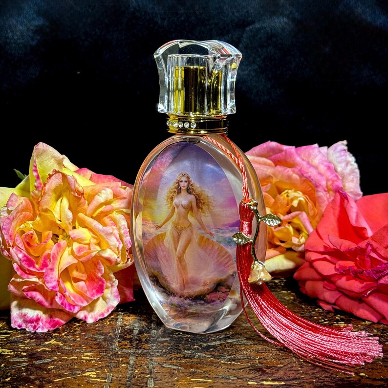 Puede incluir: Un frasco de perfume ovalado con un cuerpo de vidrio transparente, que presenta una pintura de una mujer en una concha. El frasco tiene un cuello dorado y un tap&oacute;n transparente facetado. Una borla roja con detalles dorados cuelga del frasco. Rosas en tonos rosa y amarillo rodean el frasco.