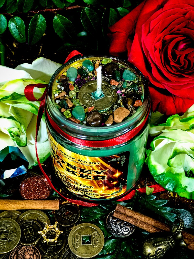 Puede incluir: Vela de vidrio verde en un frasco con una cinta roja y una vela blanca en el interior. El frasco est&aacute; lleno de piedras verdes, marrones y doradas. La etiqueta en el frasco dice "Money Maker" con las palabras "Naturally Blessed, Potently Loaded, Ritual Candle" debajo.