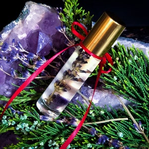 Puede incluir: Un frasco de vidrio transparente con rodillo, tapa dorada y cinta roja. El frasco est&aacute; lleno de un l&iacute;quido de color lavanda y contiene flores de lavanda secas. El frasco est&aacute; rodeado de cristales de amatista y follaje verde.