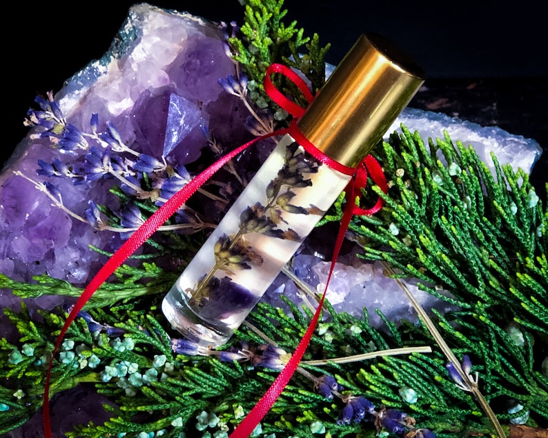 Puede incluir: Un frasco de vidrio transparente con rodillo, tapa dorada y cinta roja. El frasco est&aacute; lleno de un l&iacute;quido de color lavanda y contiene flores de lavanda secas. El frasco est&aacute; rodeado de cristales de amatista y follaje verde.