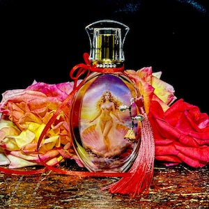 Puede incluir: Un frasco de perfume ovalado con una tapa dorada y una imagen decorativa de una mujer en una concha. La botella est&aacute; adornada con una cinta roja y una borla, y rodeada de rosas coloridas.