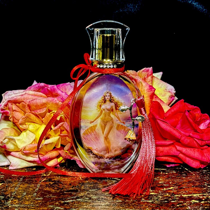 Puede incluir: Un frasco de perfume ovalado con una tapa dorada y una imagen decorativa de una mujer en una concha. La botella est&aacute; adornada con una cinta roja y una borla, y rodeada de rosas coloridas.