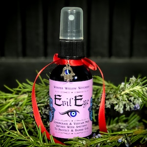 May include: A small glass bottle with a red ribbon and a silver charm. The bottle is labeled "Evil Eye" and contains a liquid. The label also says "Lemongrass & Vervain Mist Infused with Amethyst To Protect & Banish Evil."