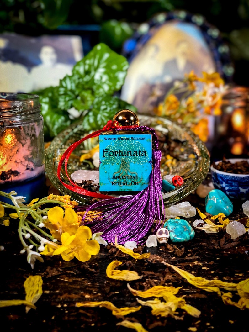 May include: A small blue bottle with a purple tassel and a red and white cord. The bottle is labeled "Fortunata Ancestral Ritual Oil" and "Winter Willow Witchery". The bottle is surrounded by yellow flowers, turquoise stones, and a skull.