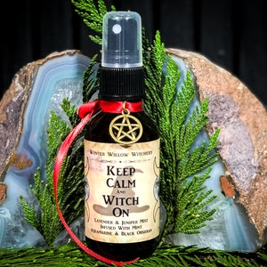 May include: A small brown glass bottle with a red ribbon and a gold pentagram charm. The bottle is labeled "Winter Willow Witchery Keep Calm and Witch On Lavender & Juniper Mist Infused with Mint Aquamarine & Black Obsidian".