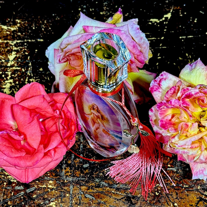 Puede incluir: Un frasco de perfume transparente con un tap&oacute;n decorativo, con una imagen pintada de una mujer. La botella est&aacute; adornada con una borla y una cinta rojas, rodeada de rosas rosas y amarillas. El fondo es una superficie oscura y texturizada.