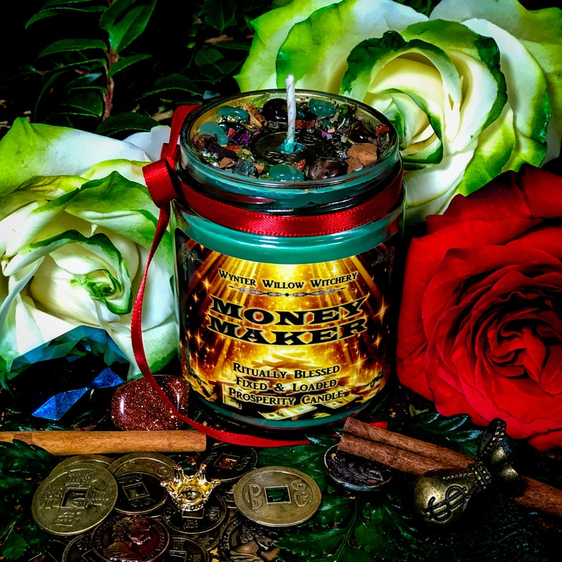 Puede incluir: Un tarro de vela de vidrio verde con una cinta roja y una etiqueta dorada que dice "Money Maker" y "Ritually Blessed Fixed & Loaded Prosperity Candle". La vela est&aacute; rodeada de rosas verdes y blancas, palitos de canela y monedas de oro.