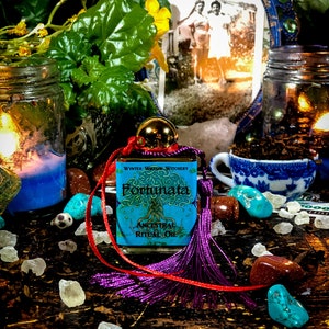 May include: A small blue bottle with a gold lid and a label that reads "Fortunata Ancestral Ritual Oil" with a purple tassel attached to a red ribbon. The bottle is sitting on a wooden surface with various crystals, stones, and a small teacup.