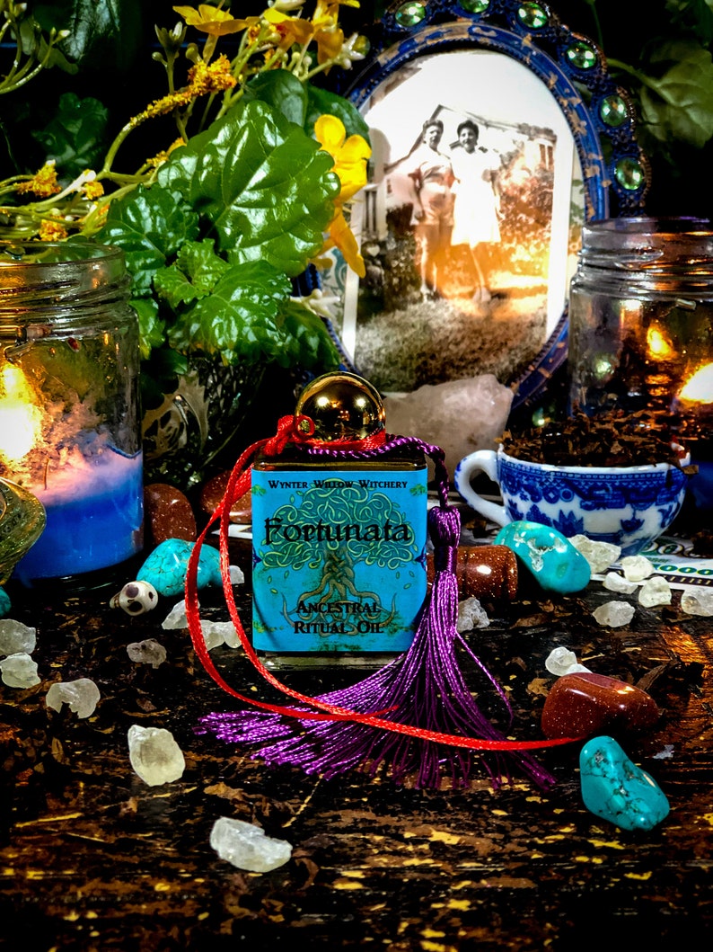 May include: A small blue bottle with a gold lid and a label that reads "Fortunata Ancestral Ritual Oil" with a purple tassel attached to a red ribbon. The bottle is sitting on a wooden surface with various crystals, stones, and a small teacup.