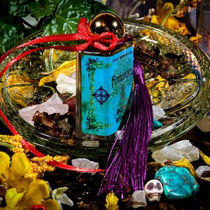 May include: A small blue glass bottle with a gold top and a purple tassel. The bottle is labeled "Fountain of Youth" and "Angelic Ritual Oil". The bottle is surrounded by crystals, dried flowers, and a skull.