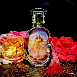 Puede incluir: Un frasco de perfume ovalado con tapa dorada y una imagen decorativa de una mujer en una concha. El frasco tiene una borla roja y est&aacute; rodeado de rosas rosas y rojas. El fondo es negro.