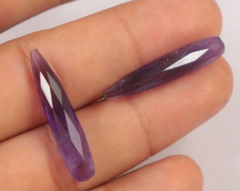 May include: Two faceted, elongated amethyst gemstones. The gemstones are a deep purple color with multiple facets that catch the light. The gemstones are teardrop shaped and appear to be ready for jewelry making.