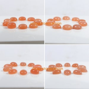 May include: A collection of 12 oval-shaped, peach-coloured gemstones. The stones are arranged in a circle on a white surface.