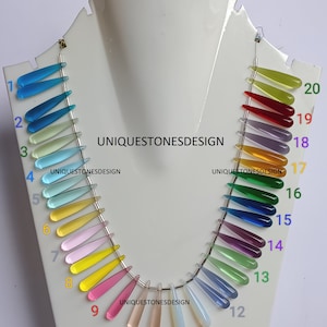 Puede incluir: Un collar con 20 cuentas de piedras preciosas en forma de lágrima en varios colores, incluyendo azul, verde, amarillo, rosa, rojo y morado. Las cuentas están enhebradas en una cadena plateada y están numeradas del 1 al 20.