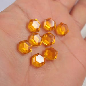 May include: A selection of amber-coloured, faceted hexagonal gemstones. The gems are scattered, highlighting their geometric form and translucent nature. The text "QUESTIONSDESIGN" is visible on one of the gems.