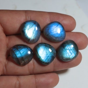 Może przedstawiać: Pięć polerowanych kamieni szlachetnych labradorytu z opalizującymi niebieskimi i turkusowymi błyskami. Kamienie są gładkie, zaokrąglone i nieznacznie różnią się kształtem. Kamienie szlachetne są wyświetlane na jasnym tle.