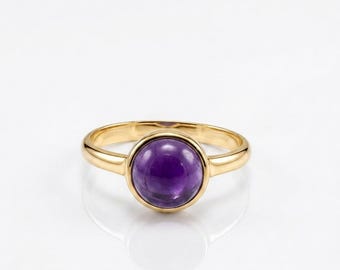 May include: A gold ring featuring a smooth, round, cabochon-cut purple amethyst gemstone set in a polished gold band. The ring has a simple, clean design with a high-shine finish, highlighting the deep violet color of the central stone.