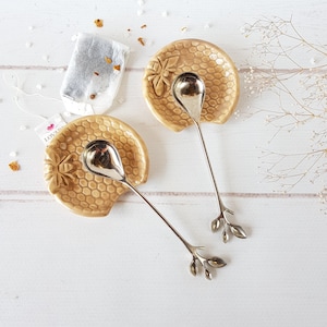 May include: Two ceramic honey comb shaped spoon rests with matching silver spoons with leaf-shaped handles. The spoon rests are beige with a raised honeycomb pattern and a bee design.