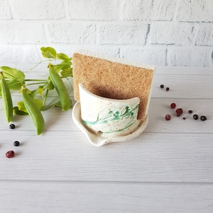 May include: White ceramic sponge holder with green and brown speckled glaze. The holder has a curved shape and is designed to hold a rectangular sponge. A brown sponge is sitting in the holder.