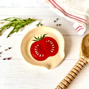 May include: A ceramic spoon rest shaped like a red tomato with a green stem. The spoon rest is on a white wooden surface.