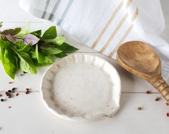 May include: A white ceramic spoon rest with a scalloped edge and a speckled glaze. The rest is sitting on a white wooden surface with a wooden spoon and green herbs.