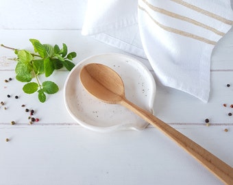 May include: White ceramic spoon rest with a speckled finish, featuring a wooden spoon resting inside. The spoon rest is on a white wooden surface with black and red peppercorns scattered around.
