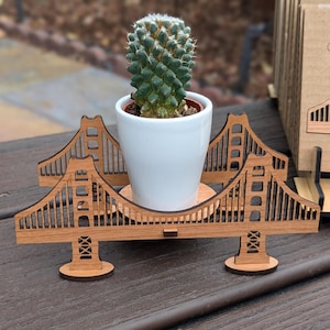 May include: A wooden Golden Gate Bridge plant stand with a small cactus plant in a white pot. The stand is made of laser-cut wood and features intricate details of the bridge.