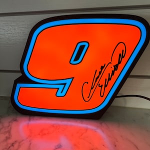Peut inclure: Un panneau lumineux en néon noir, orange et bleu en forme de chiffre 9. Le chiffre est bordé de bleu et a une bordure noire. Le chiffre est éclairé et la lumière traverse le plastique orange. Le chiffre est signé "Jeff Gordon" à l'encre noire.