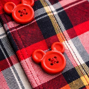 May include: Close-up of red plaid fabric with two bright red buttons. The buttons are shaped like Mickey Mouse's head. The plaid pattern includes red, black, white, and yellow lines.