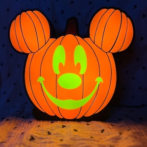 May include: A bright orange pumpkin shaped like Mickey Mouse with a green smiling face. The pumpkin is lit from inside, creating a warm glow.