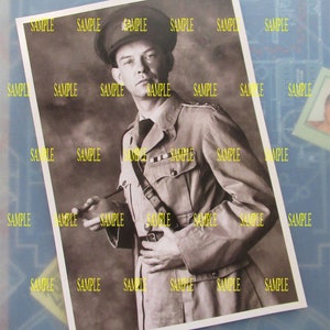 May include: A black and white photograph of a man in a military uniform. He is wearing a cap and a jacket with a belt and a strap across his chest. He is standing with his hands clasped in front of him.