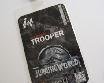 May include: A clear plastic ID badge holder with a metal clip. The badge features the text "ISLA NUBLAR TROOPER" and the "JURASSIC WORLD" logo. The background has a black and gray hexagonal pattern.