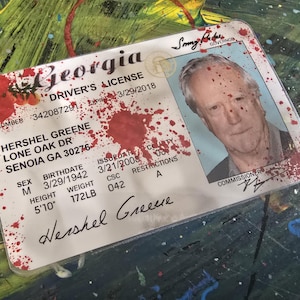 May include: A Georgia driver's license with blood splatter on it. The license is for Hershel Greene, born on 3/29/1942, with a height of 5'10" and a weight of 172 lbs. The license was issued on 3/21/2008 and expires on 3/29/2018.