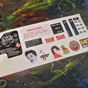 May include: A set of Ghostbusters themed stickers featuring a warning label, a character from the movie, a ghost, a monster, and a logo for Ray's Occult Books. The stickers are printed on white paper.