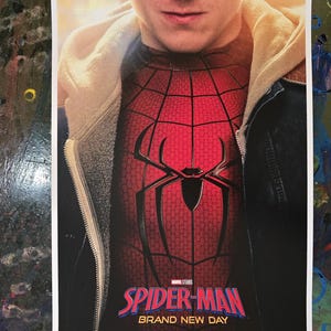 May include: A movie poster featuring a person wearing a red and black Spider-Man suit under a beige hoodie and dark jacket. The text reads Spider-Man Brand New Day, Marvel Studios, Exclusively in Movie Theatres, July 31, in Premium Formats.