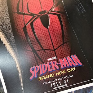 May include: A movie poster featuring a red and black textured Spider-Man suit with a large black spider emblem on the chest. The text reads Marvel Studios Spider-Man Brand New Day, Exclusively in Movie Theatres, July 31, in premium formats.