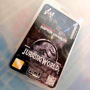 May include: A black and gray Jurassic World Raptor Handler ID card in a clear plastic sleeve. The card features the Jurassic World logo, a dinosaur graphic, and the text "ISLA NUBLAR" and "ACU".